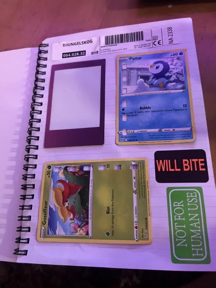 A journal page with a djungleskog tag, a blank polaroid picture, Piplup and Gossifleur pokemon cards, and veterinary labels reading 'will bite' and 'not for human use' glued to it