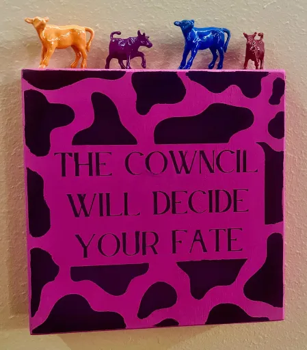 A pink sign with black cow spots and text that reads: The cowncil will decide your fate. Plastic cows in orange, purple, blue, and red stand atop the sign.