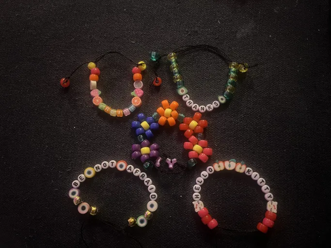 Colorful beaded bracelets. One reads 'fruity' and features multiple fruit-shaped beads. One reads 'thanks' and features avocado-shaped beads. One reads 'be not afraid' and features beads shaped like eyes. One reads 'hello kitty' and features beads shaped like hello kitty and apples. One has beads arranged into the shape of colorful flowers.