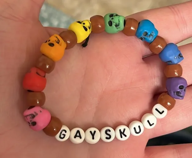 A bracelet that says 'Gayskull' and has brown pony beads and rainbow skull beads.