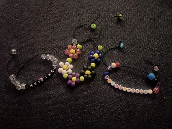 Colorful beaded bracelets. One reads 'they them' and has the genderfluid flag on it. One reads 'gender silly' and has beads in the colors of the genderfluid flag. One has beads arranged into the shape of flowers that are the colors of the genderfluid flag.